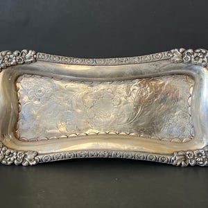 May include: Silver-toned rectangular tray with floral and scrollwork detailing. The tray has a slightly curved shape and is adorned with a floral border. The center of the tray features an engraved floral design.