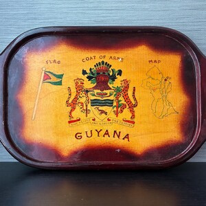May include: An oval wooden serving tray with a dark brown stained border and gold-toned metal handles featuring maroon accents. The center displays a yellow background with the Guyana coat of arms, a flag, a map, and the text "GUYANA" in black.