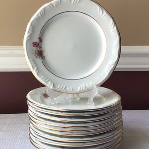 May include: A stack of ten white dinner plates with a gold trim and a floral design. The plates are stacked on a white surface.