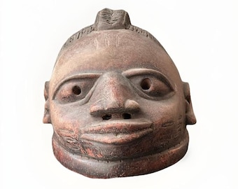 May include: A terracotta mask with a stylized human face. The mask has a textured surface and is painted in shades of brown and red.