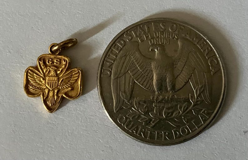 May include: A gold-colored Girl Scout pendant with the letters "GS" and an eagle emblem, next to a United States quarter coin. The coin features an eagle and the words "Quarter Dollar".