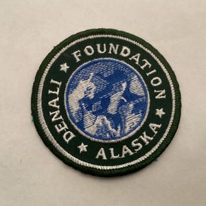 May include: Circular embroidered patch with the text "DENALI FOUNDATION ALASKA". The patch features a dark green border with white stitching and a central design in blue and white, depicting a mountain range.