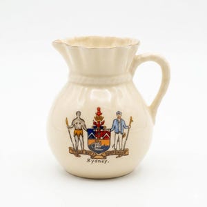 May include: A cream-colored ceramic pitcher with a ruffled rim and a single handle. The front features a colorful coat of arms depicting two figures, a ship, and a crown, with the text "I TAKE BUT I SURRENDER" and "Sydney" printed below.