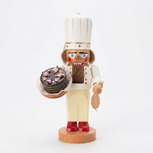 May include: A wooden nutcracker figurine dressed as a chef in a white toque and uniform, featuring brown hair and a beard. The figure holds a small, round, dark brown chocolate cake decorated with pink frosting flowers and gold accents on a tray.