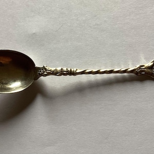 May include: A silver teaspoon with a twisted handle and a decorative design on the end. The design features a coat of arms with a building.