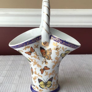 May include: White porcelain basket with a twisted handle and butterfly motif. The basket features a blue rim with gold accents and is decorated with colorful butterflies and gold floral patterns. The handle has gold stripes.