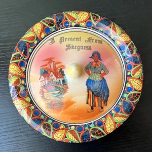 May include: A round, vintage trinket box with a colorful, patterned border. The lid features a scene with a woman in traditional clothing, a building, and the text "A Present From Skegness."