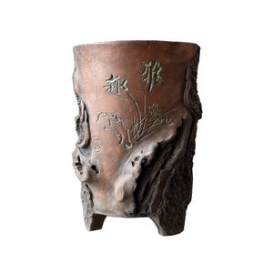 May include: A brown, three-legged planter with carved details and Chinese characters. The planter has a textured, weathered appearance, with a design of flowers and leaves. The planter is made of a dark brown material.
