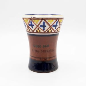 May include: A ceramic shot glass with a brown body, a navy blue base, and a decorative rim. The rim features a pattern of blue crosses, yellow, and brown accents. The text "Kero Bar Lima Sheraton" is visible on the front.