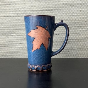 May include: A tall, blue ceramic mug with a copper-colored maple leaf design. The mug has a curved handle and a decorative base. The mug is approximately 6 inches tall.
