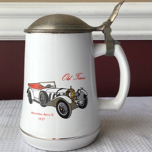 May include: White ceramic beer stein with a silver lid and handle. The stein features a vintage car illustration with the text "Old Timer" and "Mercedes-Benz S' 1927".