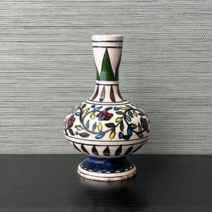 May include: A white ceramic vase with a bulbous body and a narrow neck. The vase is decorated with a colorful floral pattern in shades of red, yellow, blue, and green. The base of the vase is blue and white.