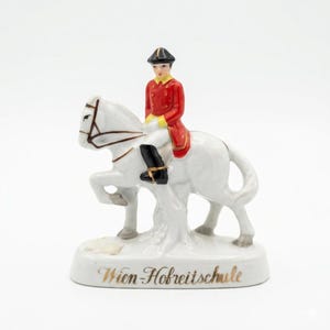 May include: A porcelain figurine of a rider on a white horse, featuring a rider in a red jacket, black hat, and black boots. The horse is in a raised leg pose. The base is inscribed with the gold text Wien-Hofreitschule in cursive script.