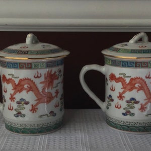 May include: Two white ceramic mugs with lids, featuring a red dragon design and gold trim. The mugs have a blue and green border with a repeating pattern.