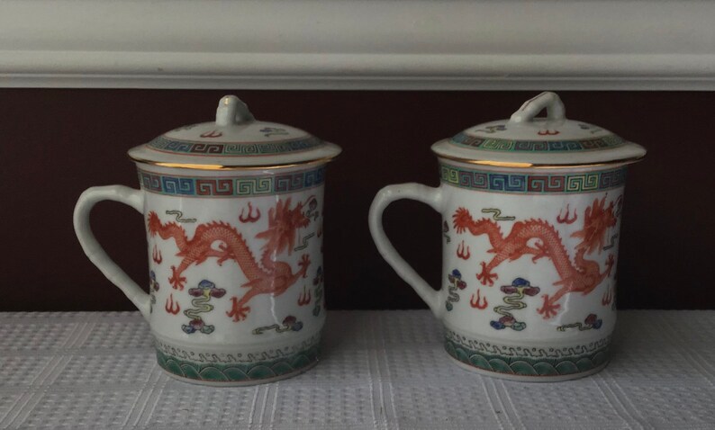 May include: Two white ceramic mugs with lids, featuring a red dragon design and gold trim. The mugs have a blue and green border with a repeating pattern.
