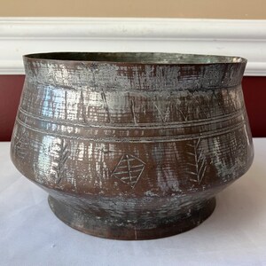 May include: A vintage copper-toned metal bowl with a wide, rounded body and a slightly flared rim. The bowl features etched geometric and leaf-like patterns around the midsection. The metal has a distressed, aged appearance with areas of lighter color.