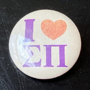 May include: A round button with the text "I ΣΠ" in purple and a peach-colored heart. The button has a white background and a glossy finish. The design is simple and bold.