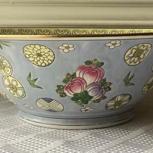 May include: A large, light blue porcelain bowl with a gold rim and a floral design. The bowl features a repeating pattern of stylized flowers, leaves, and fruit.