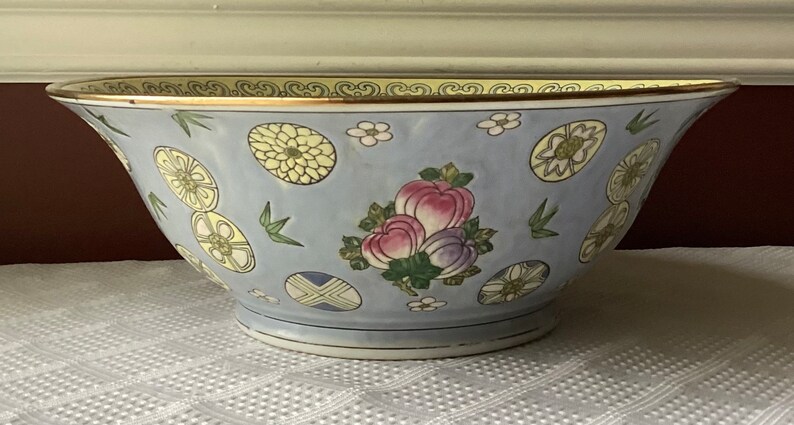 May include: A large, light blue porcelain bowl with a gold rim and a floral design. The bowl features a repeating pattern of stylized flowers, leaves, and fruit.
