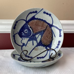 May include: Two blue and white ceramic plates with a hand-painted design of a fish. The larger plate has a brown fish with blue and white details. The smaller plate is plain white.