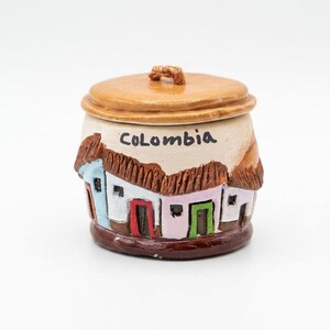 May include: A ceramic jar with a lid featuring a hand-painted scene of colorful houses with brown roofs. The word COLOMBIA is written in black letters across the top. The base is dark brown, and the lid is a smooth, light tan color with a small handle.