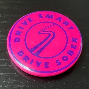 May include: A round, pink button with the words "DRIVE SMART DRIVE SOBER" in purple lettering. A purple road graphic is in the center. The button is on a dark surface.
