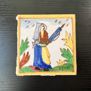 May include: A hand-painted ceramic tile depicting a woman in a blue and yellow dress holding a blue and white rolling pin. The tile has a yellow border and a white background with green and yellow floral designs.