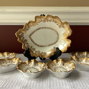 May include: A set of seven white porcelain serving dishes with gold accents. The dishes are in the shape of leaves and have a scalloped edge. The set includes a large oval dish and six smaller dishes.