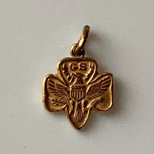 May include: A gold-colored pendant in the shape of a trefoil, featuring an eagle with outstretched wings and the letters "GS". The pendant has a loop at the top for attaching to a chain or bracelet.