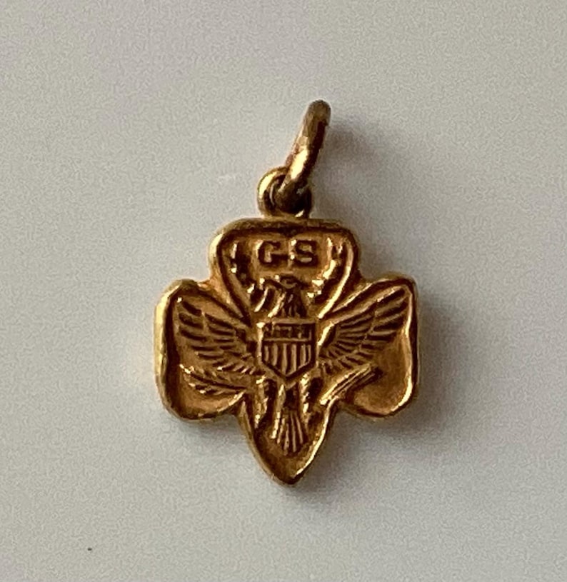 May include: A gold-colored pendant in the shape of a trefoil, featuring an eagle with outstretched wings and the letters "GS". The pendant has a loop at the top for attaching to a chain or bracelet.