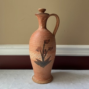 May include: A terracotta-colored ceramic pitcher with a brown floral design. The pitcher has a wide base and a narrow neck with a lid. The pitcher is approximately 12 inches tall.