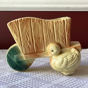 May include: A ceramic planter shaped like a wheelbarrow with a duck pulling it. The wheelbarrow is painted to look like wood and has a green wheel. The duck is yellow and has a black eye.
