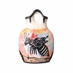 May include: A ceramic vase with a black and white bee design. The vase has a cream-colored top with a hole and a colorful background of pink, yellow, and blue. The bee is black with white stripes and is flying over a grassy area.