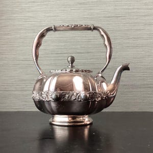 May include: A silver-colored teapot with a curved handle and spout. The teapot has a round body with decorative detailing around the base and lid. The handle is arched and attached to the top of the pot.