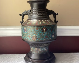 May include: A large, antique bronze vase with intricate cloisonné enamel decoration. The vase features a turquoise blue background with floral patterns in gold, red, and white. The vase has two elephant head handles and sits on a wooden base.