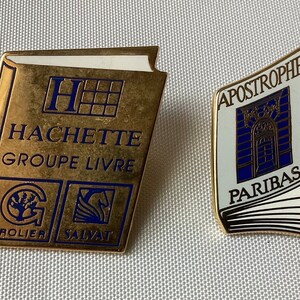 Op de afbeelding: Twee gouden boekvormige pinnen. De eerste pin heeft de tekst "HACHETTE GROUPE LIVRE" en de logo's "GROLIER" en "SALVAT". De tweede pin heeft de tekst "APOSTROPHES PARIBAS" en een blauwe afbeelding van een gebouw.