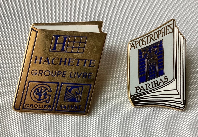 Op de afbeelding: Twee gouden boekvormige pinnen. De eerste pin heeft de tekst "HACHETTE GROUPE LIVRE" en de logo's "GROLIER" en "SALVAT". De tweede pin heeft de tekst "APOSTROPHES PARIBAS" en een blauwe afbeelding van een gebouw.