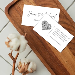 May include: White wedding invitation card with black text that reads "Jenn ♥ Michael" and "AUGUST". A black heart-shaped QR code is on the bottom right corner of the card. The text below the QR code reads "For more wedding details and to RSVP on our wedding website, please scan this QR code or visit our wedding website at www.jennandmichael.com".