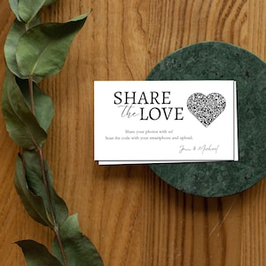 May include: White card with black text that reads "SHARE the LOVE". A black and white QR code in the shape of a heart is on the card. The text below the QR code reads "Share your photos with us! Scan the code with your smartphone and upload. Jenn & Michael"