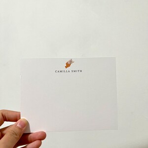 Op de afbeelding: Een witte kaart met een kleine oranje en witte illustratie van een schelp bovenaan. De tekst "CAMILLA SMITH" is in zwart onder de illustratie gedrukt.