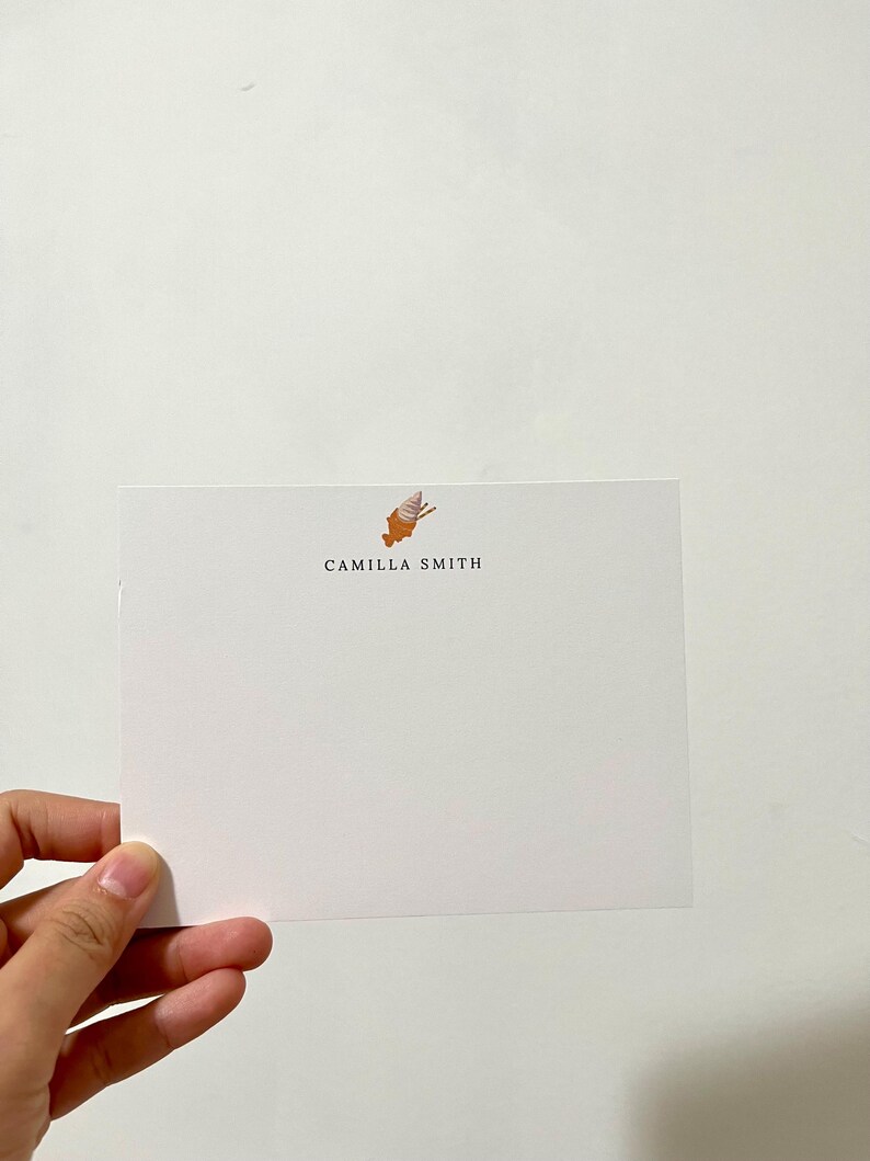 Op de afbeelding: Een witte kaart met een kleine oranje en witte illustratie van een schelp bovenaan. De tekst "CAMILLA SMITH" is in zwart onder de illustratie gedrukt.