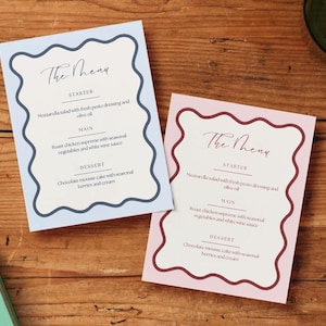 May include: Two menu cards with a wavy border. One is light blue, the other is pink. Both cards have the text "The Menu" at the top, followed by a list of dishes: starter, main, and dessert. The cards are on a wooden surface.