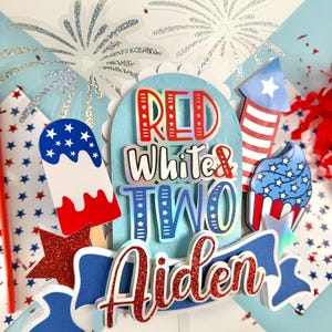 May include: Red, white, and blue cake topper with the words "Red White & Two Aiden" and a red glitter star. The topper features a red, white, and blue popsicle, cupcake, and firework.