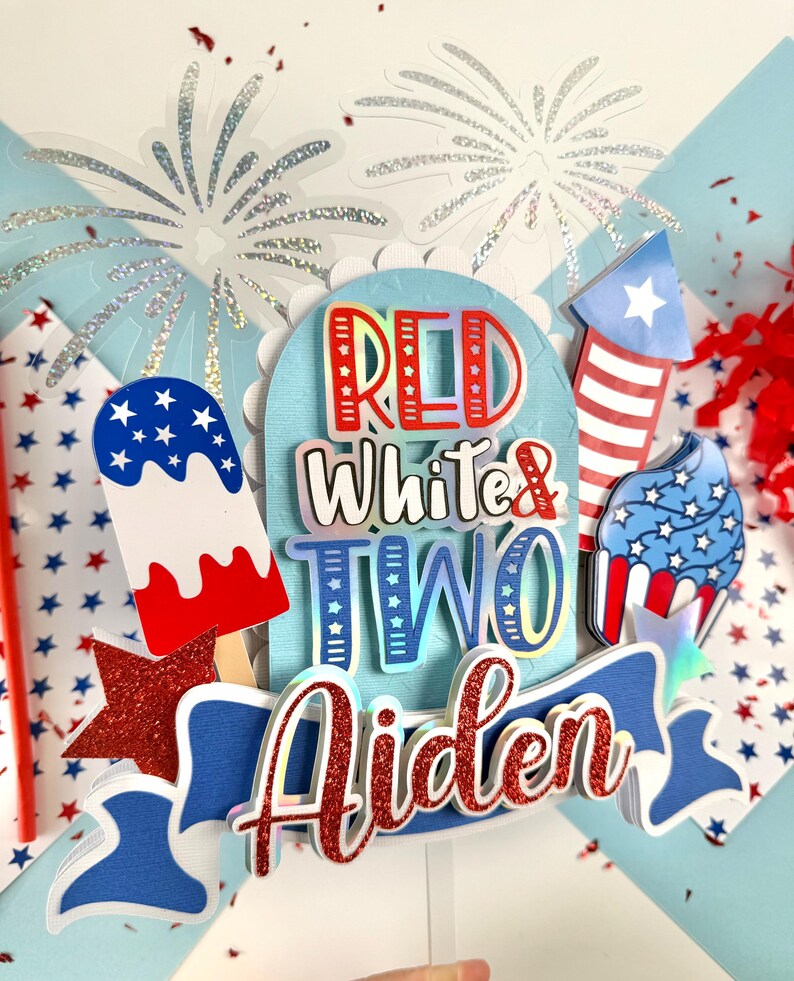 May include: Red, white, and blue cake topper with the words "Red White & Two Aiden" and a red glitter star. The topper features a red, white, and blue popsicle, cupcake, and firework.