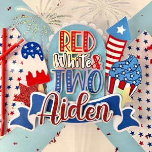 May include: Red, white, and blue cake topper with the words "Red White & Two Aiden" and a firework, cupcake, and popsicle design.