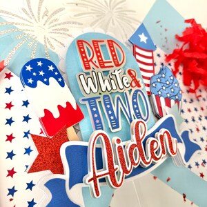 May include: Red, white, and blue cake topper with the words "Red White & Two" and the name "Aiden" in red glitter. The topper also features a firework, an ice cream cone, and a cupcake.