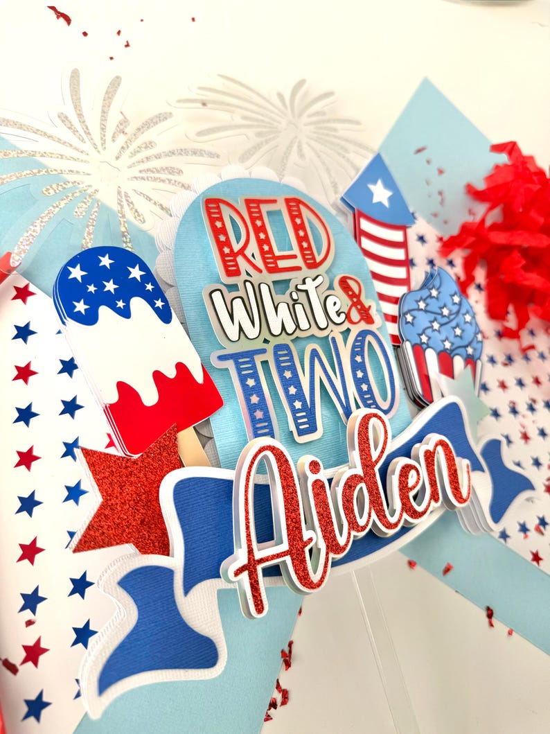 May include: Red, white, and blue cake topper with the words "Red White & Two" and the name "Aiden" in red glitter. The topper also features a firework, an ice cream cone, and a cupcake.