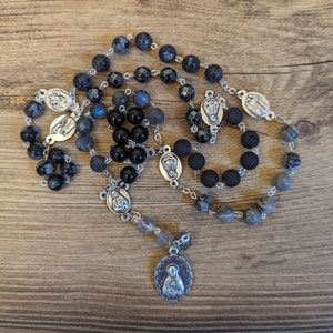May include: A black and grey rosary with silver beads and charms. The rosary features a silver pendant with a depiction of the Virgin Mary.