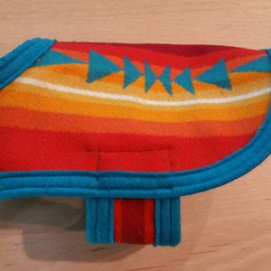 May include: A turquoise blue and red dog sweater with a southwestern pattern. The sweater has a turquoise blue trim and a red and turquoise blue strap.