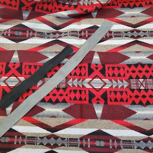 Pode incluir: Um tecido estampado em vermelho, preto e cinza, com um design geométrico de diamantes e triângulos. Existem duas tiras de material: uma preta, uma cinza. O padrão do tecido é intrincado.